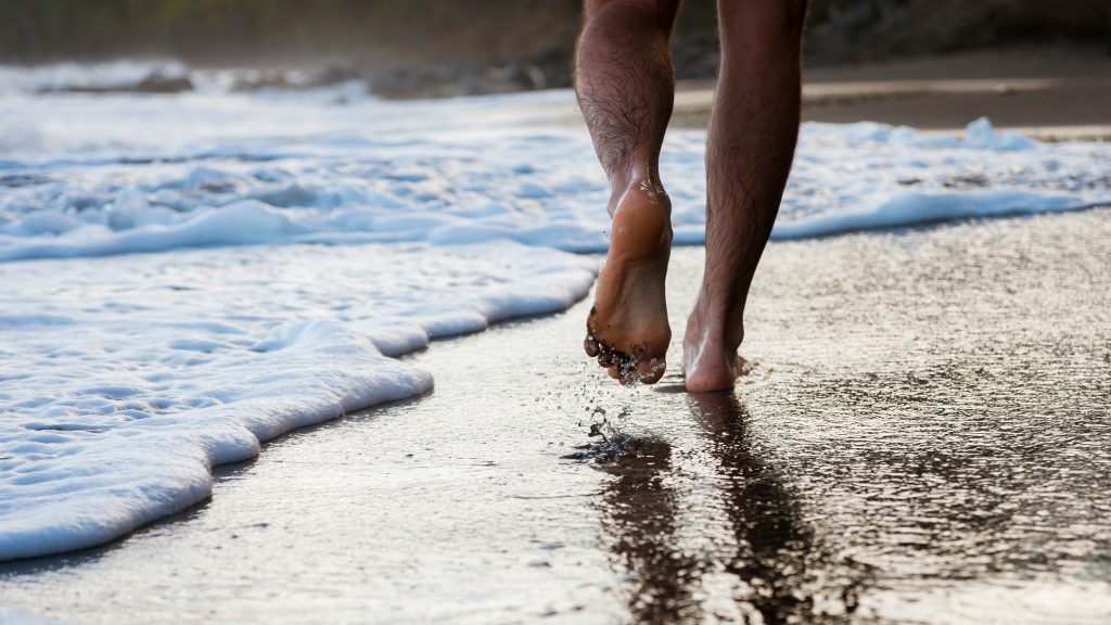 Ein Fuß wird Richtung Kamera gezeigt während ein Mann am Strand entlangläuft. Man sieht Wellen und Sand an den Zehen