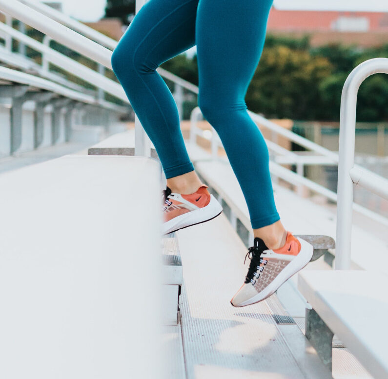 Frau in einem weißen Sporttop, in türkizen Sportleggings und orangenen Sportschuhen läuft weiße Treppen hoch Seitenansicht
